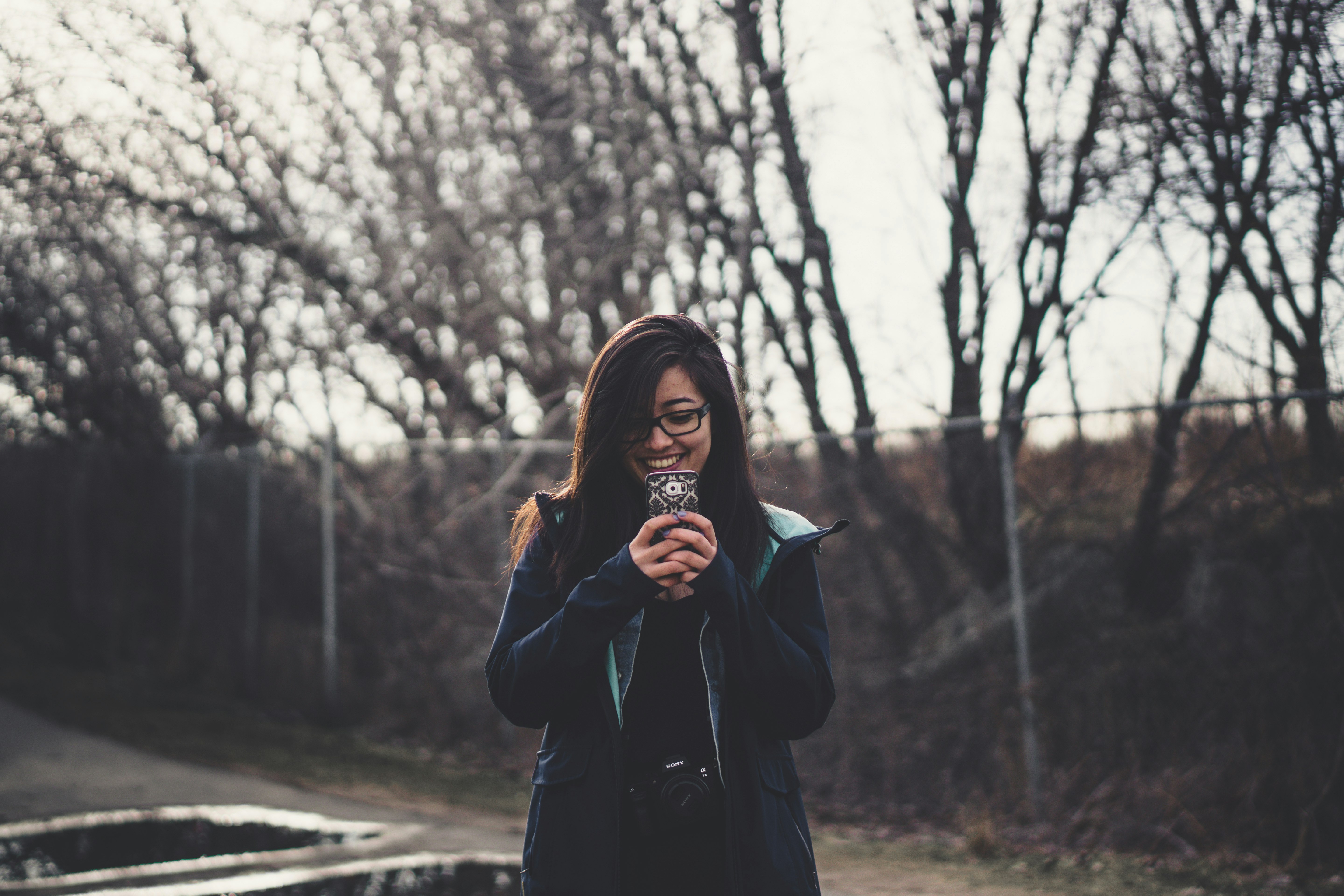 smiling woman holding smartphone standing near fence with trees during daytime